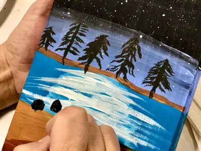 Close up of a hand painting a scene on a wooden paddle