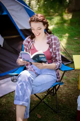 A woman sits in a lawn chair reading a book in front of a blue tent
